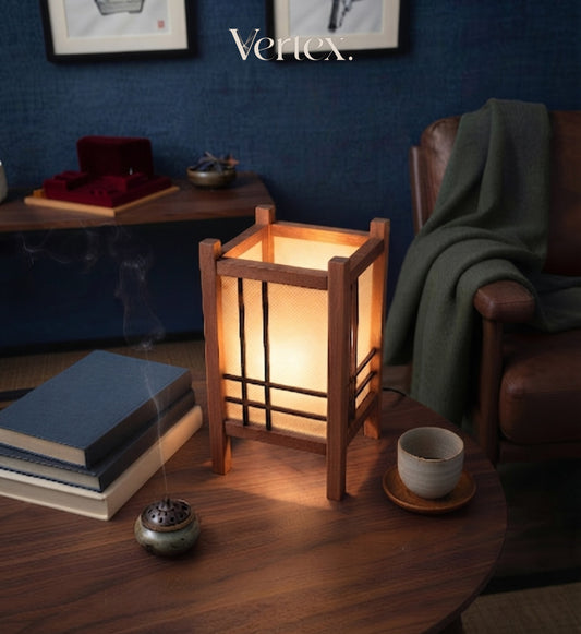 A warm, glowing Japanese-style wooden lantern with a white paper screen sits on a dark wooden table in a cozy, dimly lit room. A green throw blanket is draped over a brown leather armchair in the background, and a book, incense burner, and ceramic cup are on the table.