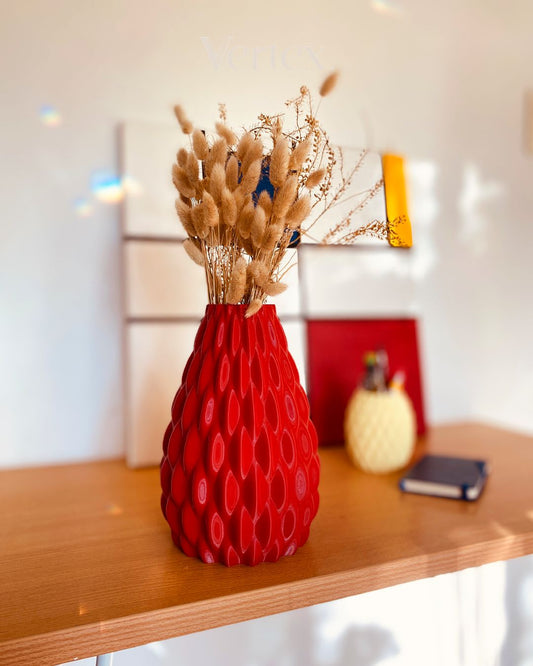 A textured red vase filled with dried bunny tail grass, displayed on a wooden shelf in a brightly lit room.