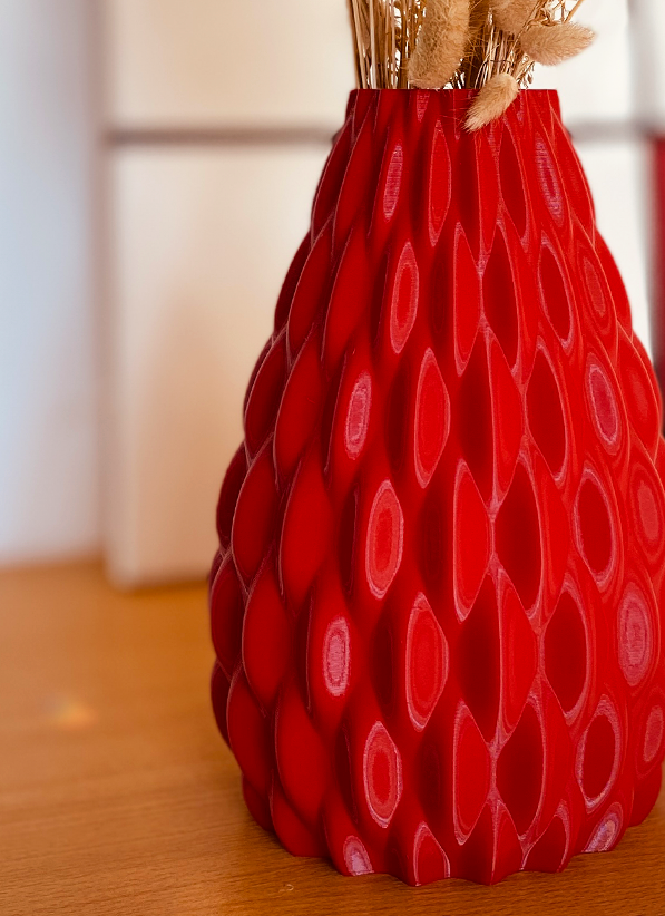 A textured red ceramic vase filled with dried bunny tail grass, displayed on a wooden shelf in a modern, softly lit room.