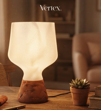 A modern table lamp with a textured white frosted shade and a terracotta-colored sculptural base, shown lit on a wooden table in a warm, softly blurred interior setting. The word 'Vertex.