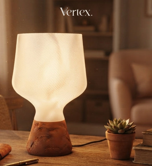 A modern table lamp with a textured white frosted shade and a terracotta-colored sculptural base, shown lit on a wooden table in a warm, softly blurred interior setting. The word 'Vertex.
