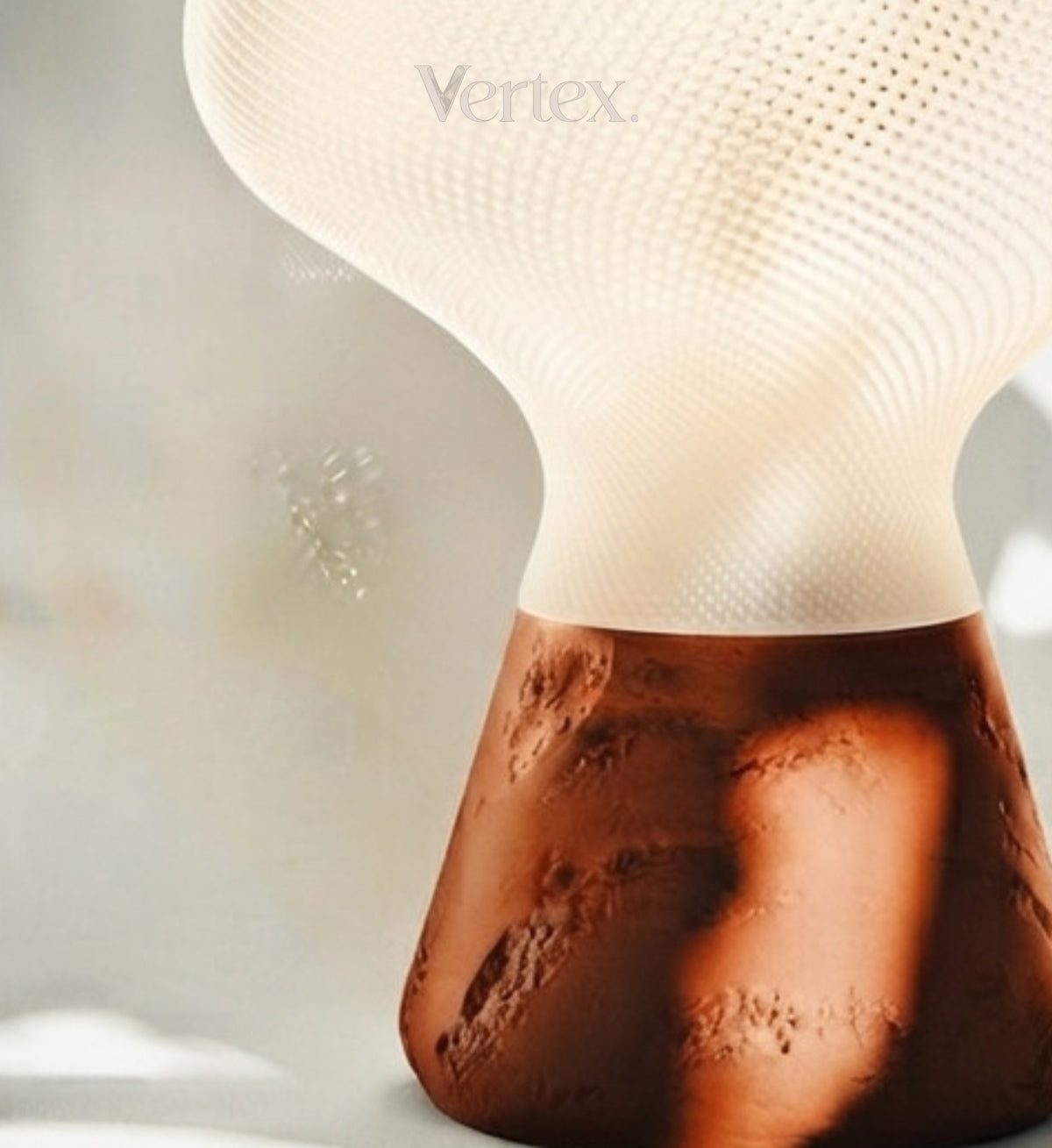 A close-up of a modern table lamp with a textured white upper shade and a sculptural, cratered brown base, featuring the Vertex brand name.
