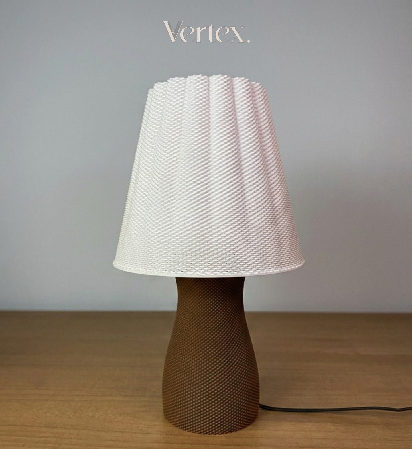 A table lamp with a textured white pleated shade and a brown textured base, sitting on a wooden surface against a gray wall. The brand name 'Vertex.