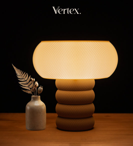 A textured table lamp with a glowing warm white dome shade and a ribbed brown base, displayed on a wooden surface next to a small vase with dried leaves. The brand name 'Vertex.
